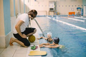 A man kneeling down next to two children in the pool.